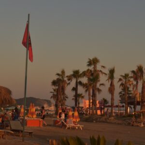 חוף בדורס בשקיעה, עם עצי דקל, שמשיות קש ואנשים נהנים מהערב